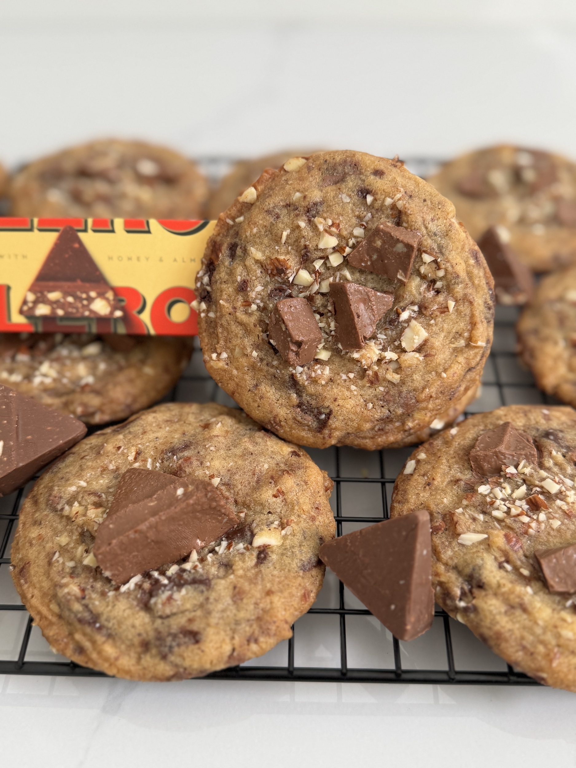Brown Butter Almond Honey Toblerone Cookies piled on a cooling sheet with a single cookie propped up by a Toblerone package